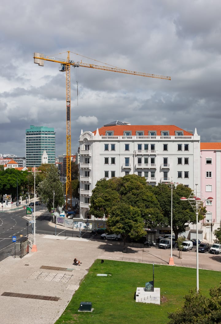 A tall yellow construction crane rising over a white apartment building in Lisbon, Portugal, with the city skyline behind under a cloudy sky