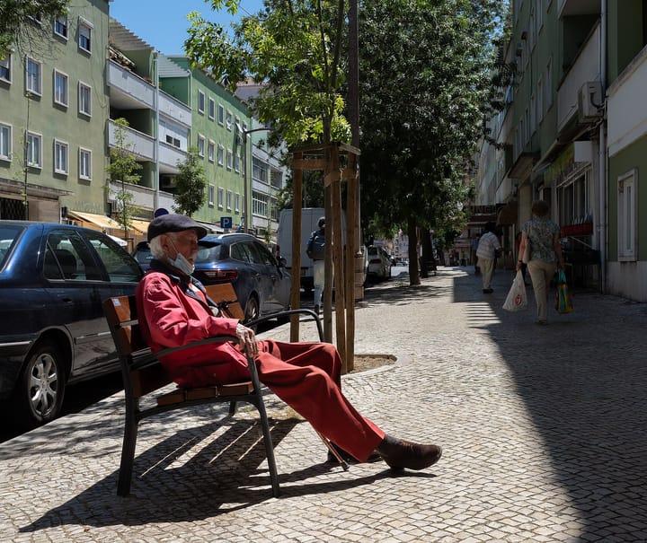 An elderly man in a red jacket and flat cap resting on a bench on a cobbled Lisbon street