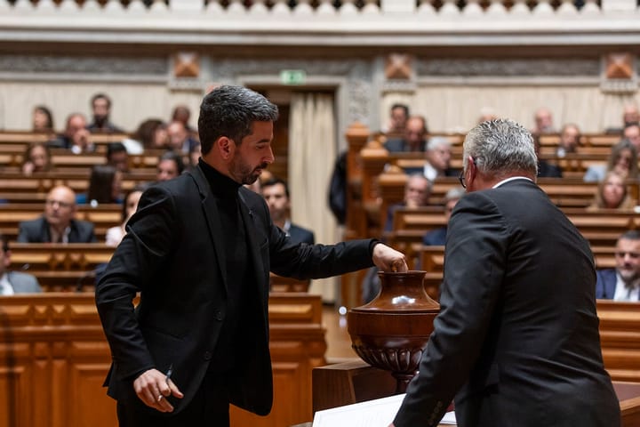 A deputy places a ballot into a tall wooden urn during a plenary vote in the chamber of the Assembleia da Republica in Lisbon, with rows of seated members behind