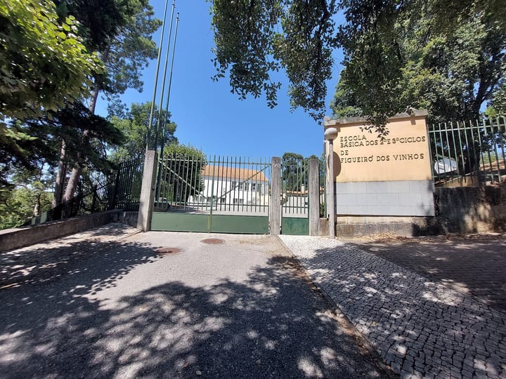 The green gated entrance of the Escola Basica dos 2.o e 3.o Ciclos de Figueiro dos Vinhos, its name carved into a stone wall beside the gate, with the white school building behind