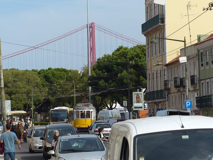 A busy Lisbon street clogged with cars and a yellow tram, with the red 25 de Abril bridge rising in the background