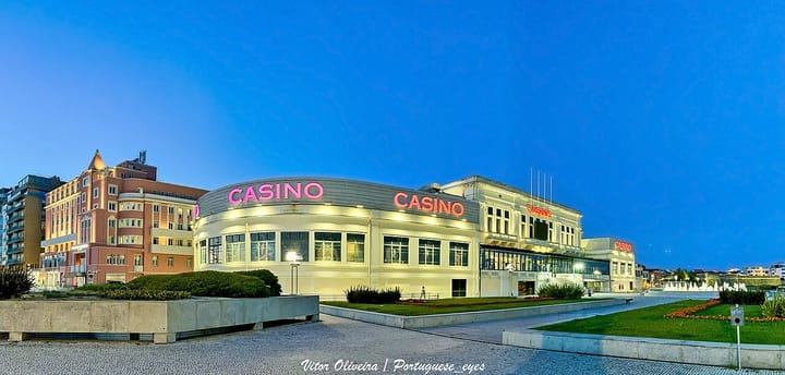 The Casino da Póvoa in Póvoa de Varzim lit up at dusk, its cream facade glowing with pink neon CASINO signage under a deep blue sky