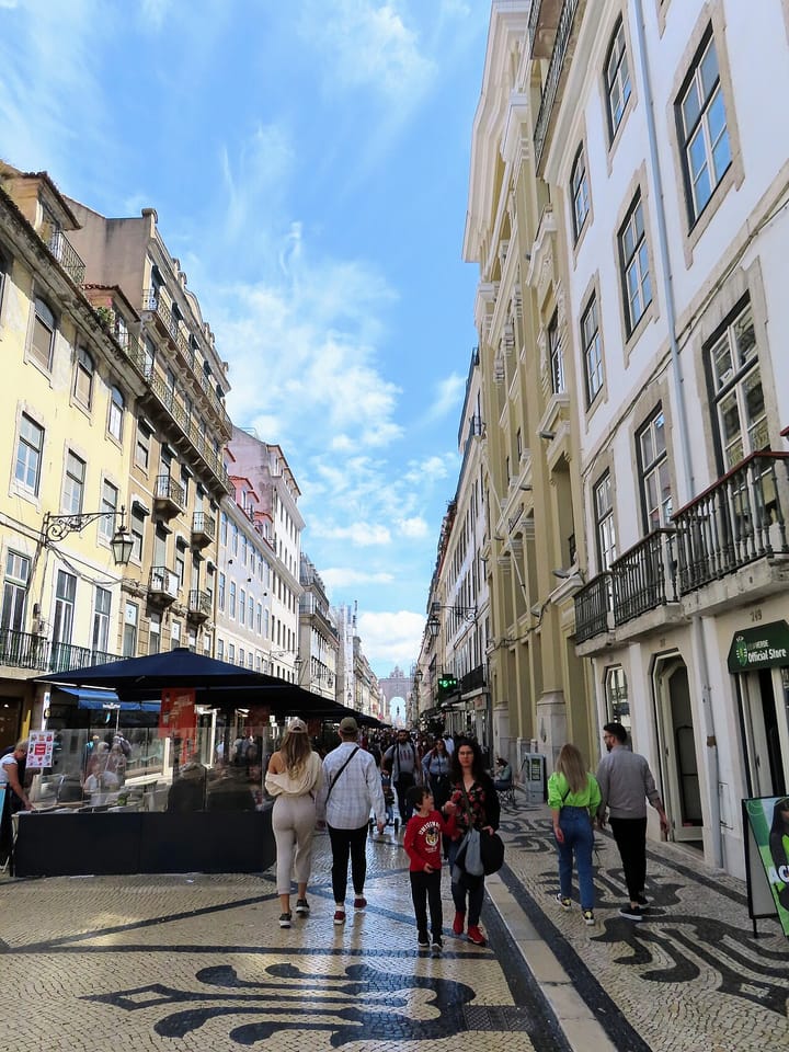 Pedestrians walking along Rua Augusta, Lisbon's main downtown pedestrian street, on the black-and-white calçada pavement between tall buildings, with the Rua Augusta arch in the distance