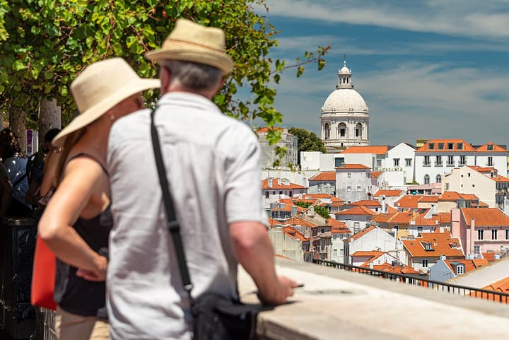 Two visitors in sun hats look out over the terracotta rooftops of Lisbon's Alfama district toward the dome of the National Pantheon