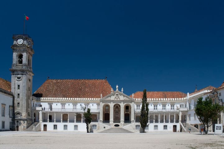 The Paco das Escolas courtyard of the University of Coimbra, with its baroque clock tower flying the Portuguese flag and the white Rectory building under a blue sky