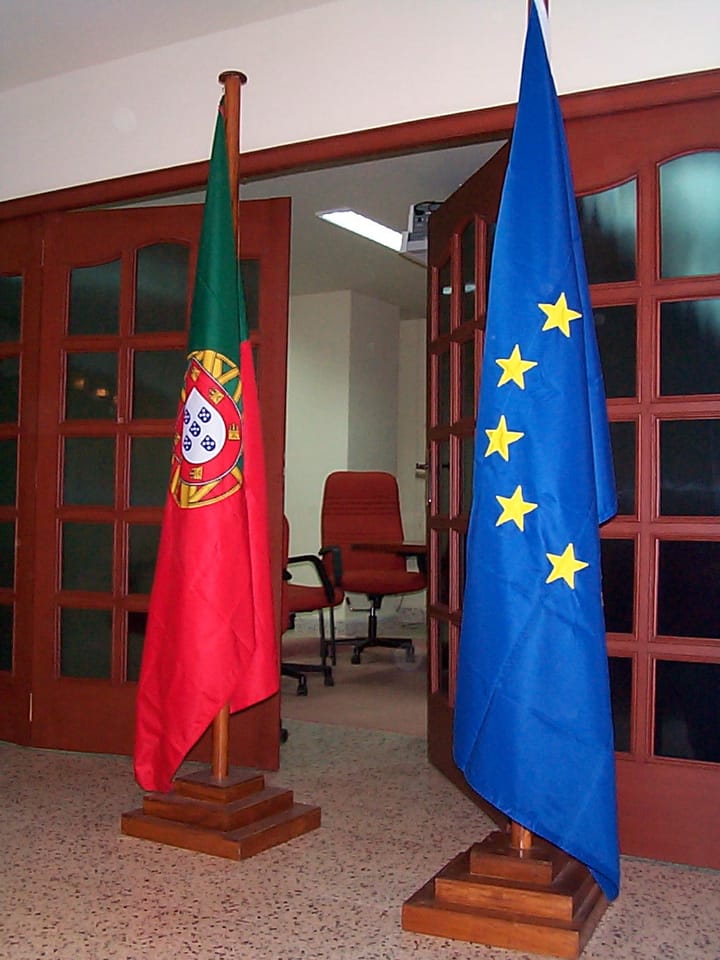 The flag of Portugal and the flag of the European Union standing on wooden poles beside an open doorway in an office