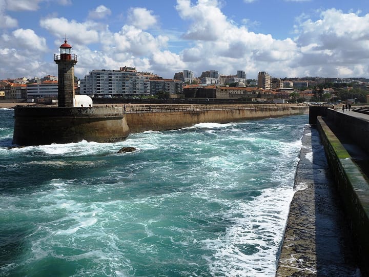 The Felgueiras lighthouse on its stone pier at Foz do Douro in Porto, with waves breaking around it and the city's seafront behind under a blue sky