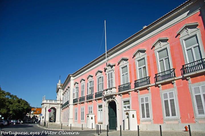 The long pink facade of the Palacio das Necessidades in Lisbon, seat of Portugal's foreign ministry, with white sentry boxes flanking the main door and a bare flagpole above the roofline.