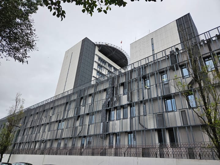 The grey metal and glass frontage of the Policia Judiciaria national headquarters in Lisbon, with a taller office tower and its curved roof canopy rising behind.
