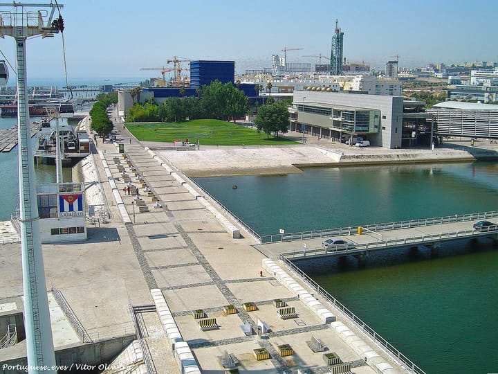 The Parque das Nacoes waterfront in Lisbon seen from above, a cable car pylon in the foreground, a dock and footbridge, office blocks and construction cranes on the skyline.