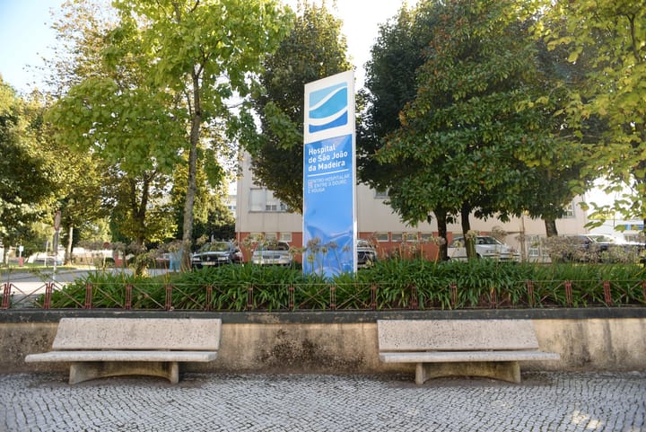 A tall blue sign reading Hospital de São João da Madeira, Centro Hospitalar de Entre o Douro e Vouga, standing in a landscaped square in front of the hospital building between trees and two s