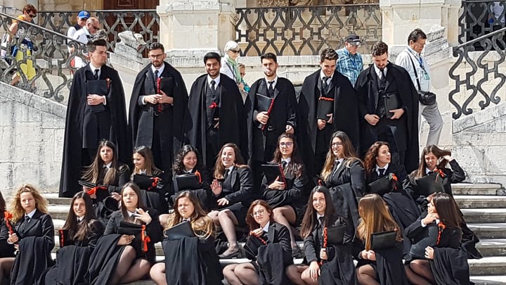 A group of Coimbra university students in the traditional black capes and gowns of the traje académico sit and stand together on the stone steps of the university