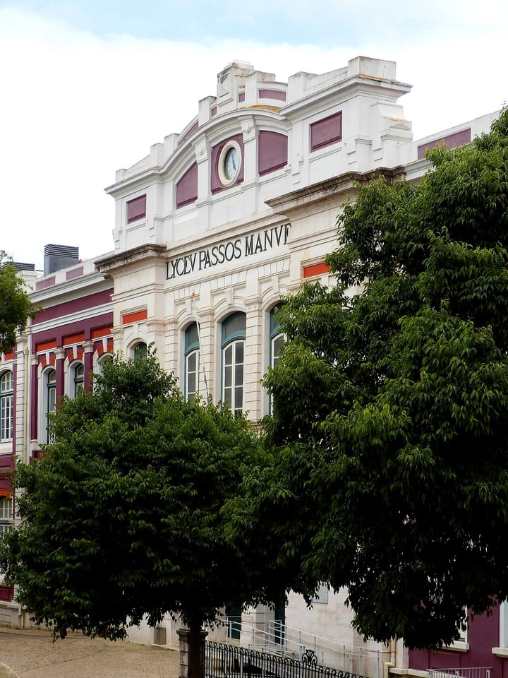 The white and burgundy facade of the Liceu Passos Manuel in Lisbon, lettered LYCEV PASSOS MANVEL beneath a clock set in the pediment, seen over street trees.