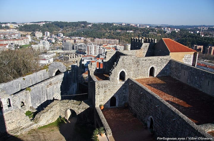 The stone walls and tiled roofs of Leiria castle in the foreground, with the city's apartment blocks and the wooded Lis valley spread out behind under a clear sky.