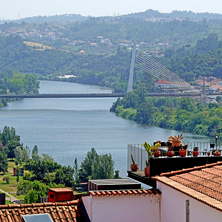 The River Mondego flowing past the terracotta rooftops of Coimbra beneath a white cable-stayed bridge, with wooded hills rising behind