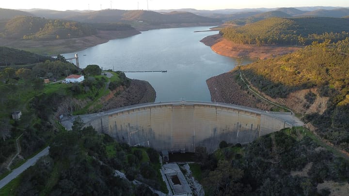 An aerial view of the curved concrete Bravura dam and its reservoir among wooded Algarve hills near Lagos, with a pale drawdown ring along the far bank.