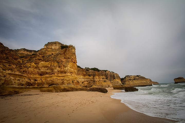 The golden limestone cliffs and sandy shore of Praia da Marinha in Lagoa, Algarve, under a grey sky, illustrating the region's sun-and-sand tourism
