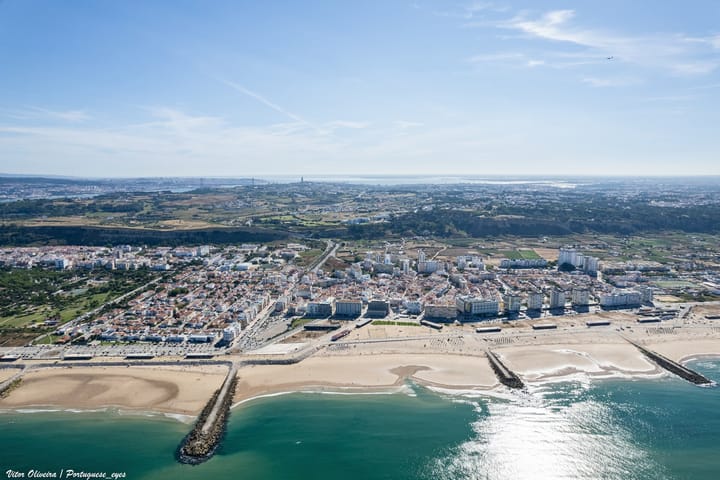 An aerial view of Costa da Caparica, the town's white blocks and red roofs behind a wide sand beach with rock groynes, and Lisbon and the 25 de Abril bridge on the far horizon.