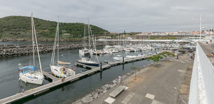Yachts moored at the marina of Angra do Heroísmo on Terceira in the Azores, with the wooded slope of Monte Brasil and the town's white buildings behind under an overcast sky.