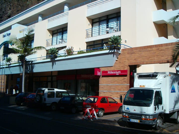 A Millennium bcp bank branch with its red sign on a Portuguese street, with cars and a delivery van parked outside in the sun