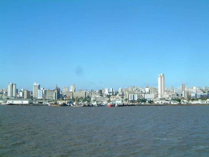 The Maputo waterfront seen from the water, cargo vessels and cranes along the quay with the city's mid-rise skyline and a tall white tower behind under a clear blue sky.