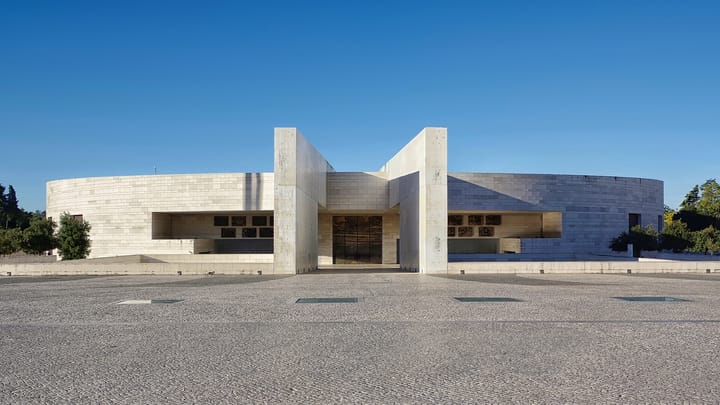 The Basílica da Santíssima Trindade at the Sanctuary of Fátima, a low circular stone building with a wide central entrance, seen across the empty paved esplanade under a clear blue sky.