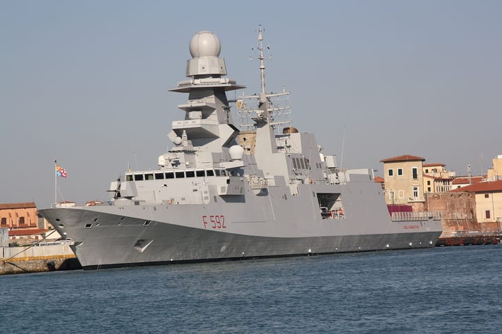 The Italian Navy FREMM frigate Carlo Margottini, pennant number F 592, moored bow-on at a harbour quay with its grey superstructure and radar dome against a clear sky.
