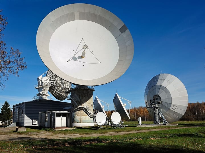 A satellite ground station with one large white parabolic antenna and a field of smaller dishes on a grassy site under a blue sky