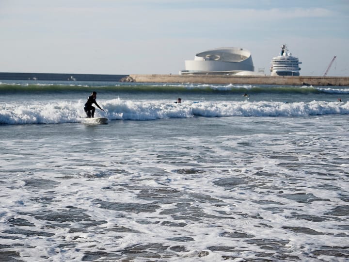A surfer riding whitewater at Matosinhos beach near Porto, with the Leixões breakwater and the spiral white cruise terminal building and a moored cruise ship on the horizon behind.