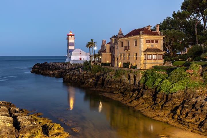 The Santa Marta lighthouse and the Condes de Castro Guimaraes house on the rocky Cascais shoreline at dusk, lit windows reflected in the shallow water below.