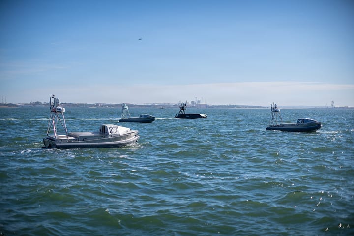 Four small grey unmanned surface vessels under way on the Sado estuary off Setubal, each with a mast-mounted radar and camera pod, the port's industrial shoreline behind.