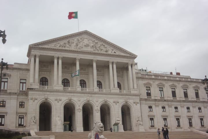 The neoclassical facade of the Palacio de Sao Bento in Lisbon, seat of the Portuguese parliament, with the national flag above the pediment and a stone lion at the foot of the steps.