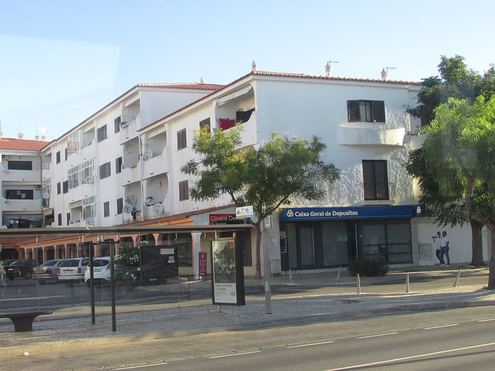 A Caixa Geral de Depositos bank branch with its blue sign on the ground floor of white Portuguese apartment blocks in Albufeira, seen from across the road with a bus shelter in front