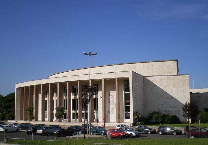 The long colonnaded limestone facade of the Reitoria da Universidade de Lisboa on the Cidade Universitaria campus under a clear blue sky, with cars parked along the road in front