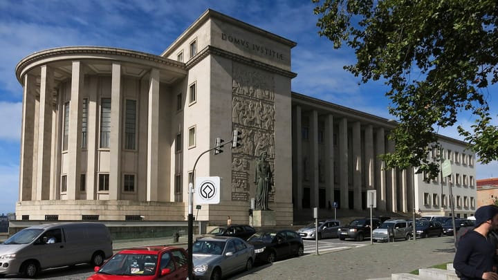 The stone DOMVS IVSTITIAE facade and colonnade of the Palacio da Justica courthouse in Porto, Portugal, with a carved relief and a statue beside the entrance.