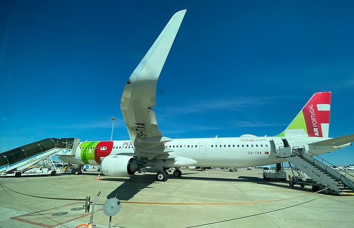 A white TAP Air Portugal Airbus jet in green-and-red livery reading 'AIR PORTUGAL', parked at a boarding gate at Lisbon Airport under a clear blue sky