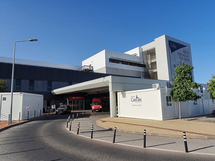 The main entrance and emergency bay of the Hospital de Cascais Dr. Jose de Almeida, a modern Portuguese public hospital, with an ambulance parked outside under a clear blue sky