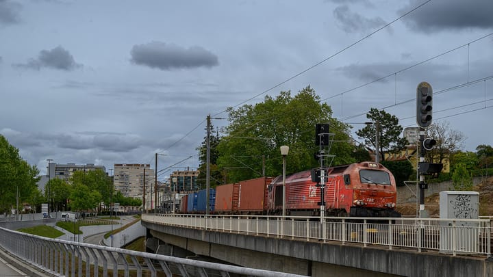 A red Takargo container freight train crosses a viaduct through a Portuguese city under a grey sky, illustrating the rail-freight focus of the planned Grandola logistics park