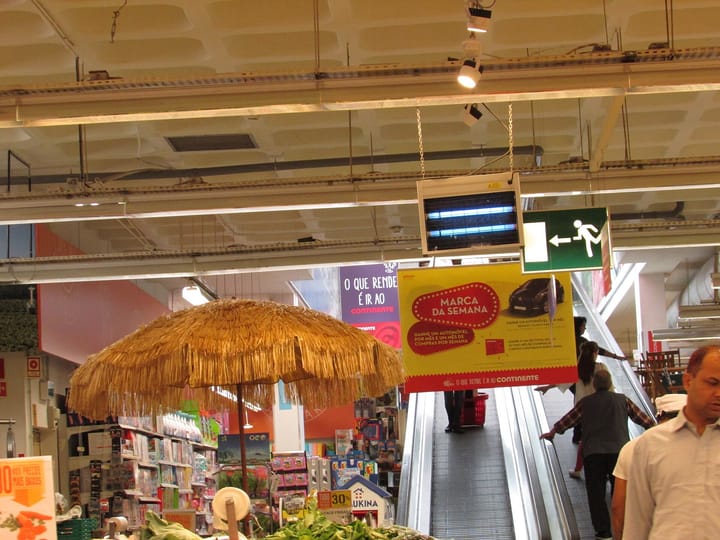 The interior of a Continente supermarket in Portugal, with fresh produce in the foreground and a promotional sign above the escalator, illustrating consumer prices and the cost of living