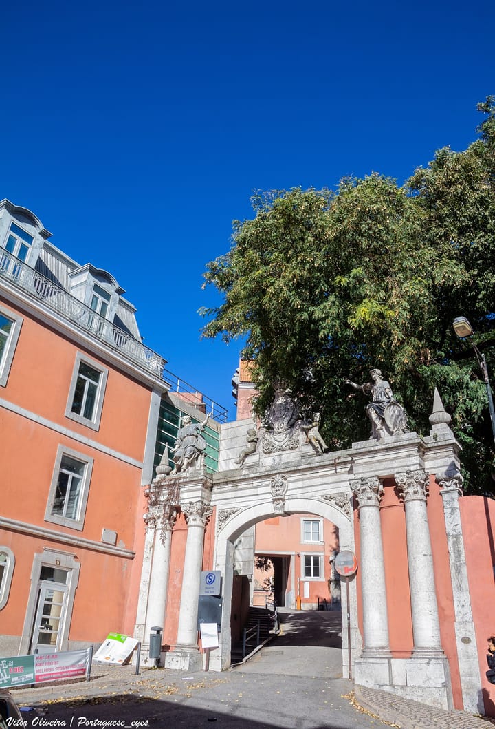 The baroque stone gateway of the Hospital de São José in Lisbon, with statues and a coat of arms above the arch, a hospital signboard beside it and a trade union banner on the pavement.