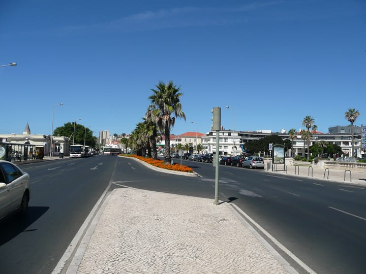 The Avenida Marginal dual carriageway at Estoril, with palm trees on the central reservation, buses at a stop and the seafront buildings of Cascais beyond