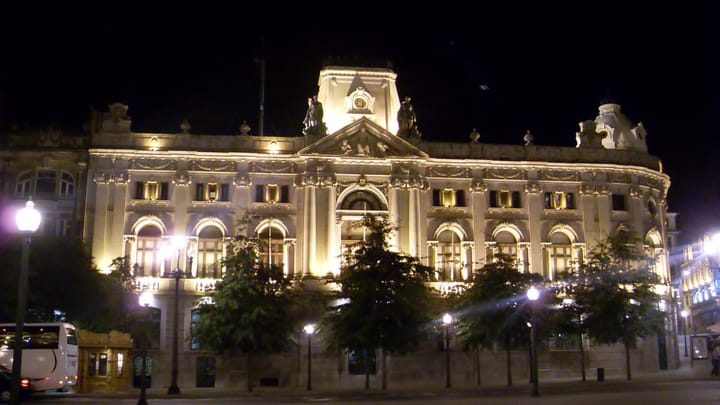 The floodlit neoclassical Banco de Portugal building on Avenida dos Aliados in Porto at night, its columned facade and central clock tower lit from below behind a row of trees