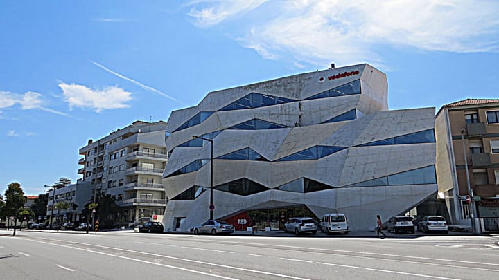The angular concrete and glass Edificio Vodafone on Avenida da Boavista in Porto, its folded facade lit by sun under a blue sky, with parked cars and an empty road in the foreground