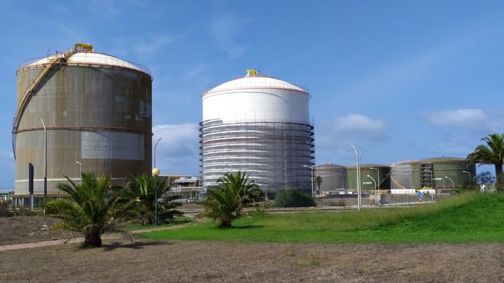 Two huge cylindrical storage tanks, one weathered steel and one white and scaffolded, at the liquefied natural gas terminal in Sines, with smaller green tanks and palms under a blue sky