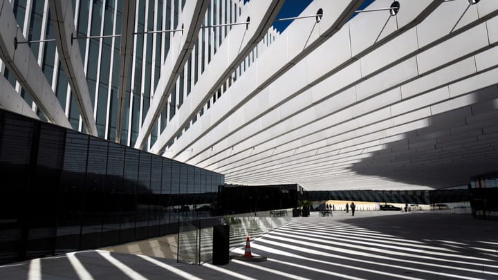 Sunlight striping the paving beneath the long white concrete colonnade of the EDP headquarters in Lisbon, with a dark glass wing on the left and a few people walking in the distance