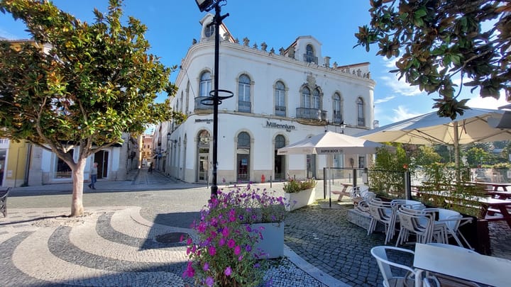 A white corner building with arched windows and iron balconies housing a Millennium bcp branch on a cobbled Leiria square, magnolia trees and a sunlit cafe terrace on either side