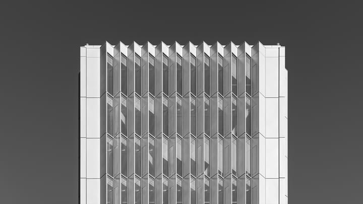 Black and white photograph of the upper floors of a modern Lisbon office tower, its glass facade broken into narrow angled vertical fins, set against a plain dark sky