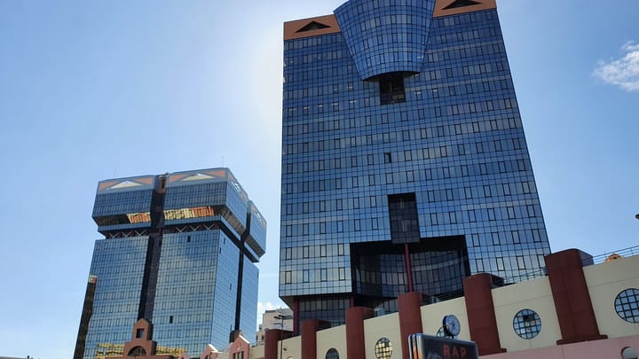 Two blue mirrored-glass postmodern office towers with terracotta trim, the Torres das Amoreiras, rising above a pink shopping centre facade against a clear sky in Lisbon