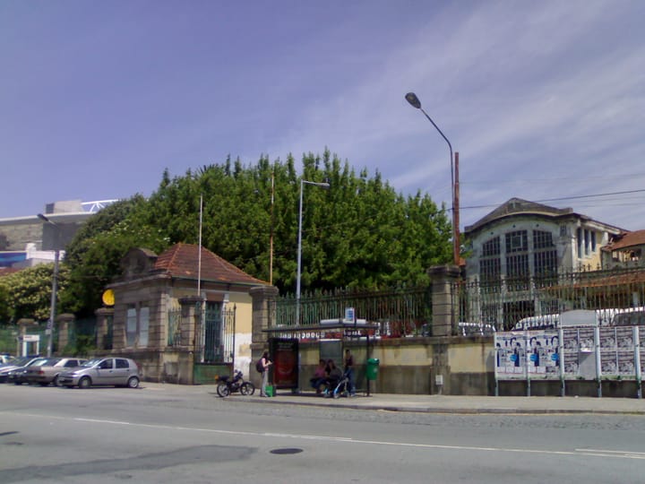 The gate and derelict pavilion of the old Matadouro Industrial do Porto beside the road at Campanha, with the roofline of the Estadio do Dragao visible behind the trees.
