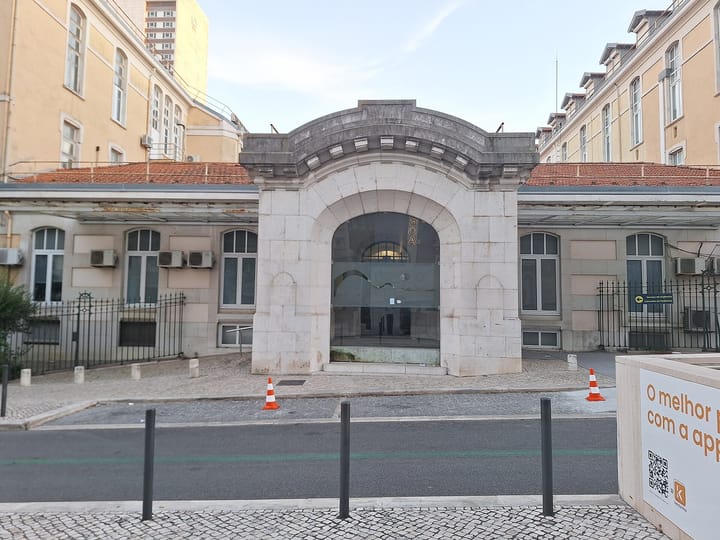The arched stone entrance of the Maternidade Alfredo da Costa in Lisbon, seen from Rua Pinheiro Chagas, with a sign for the emergency department and paediatric clinic beside the gate.